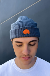 Person wearing classic cuffed knit fisherman beanie featuring embroidered Rooted Swim sunset logo in a blue gray color. high quality knit beanie
