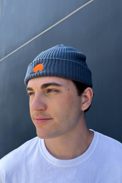 Person wearing classic cuffed knit fisherman beanie featuring embroidered Rooted Swim sunset logo in a blue gray color. high quality knit beanie