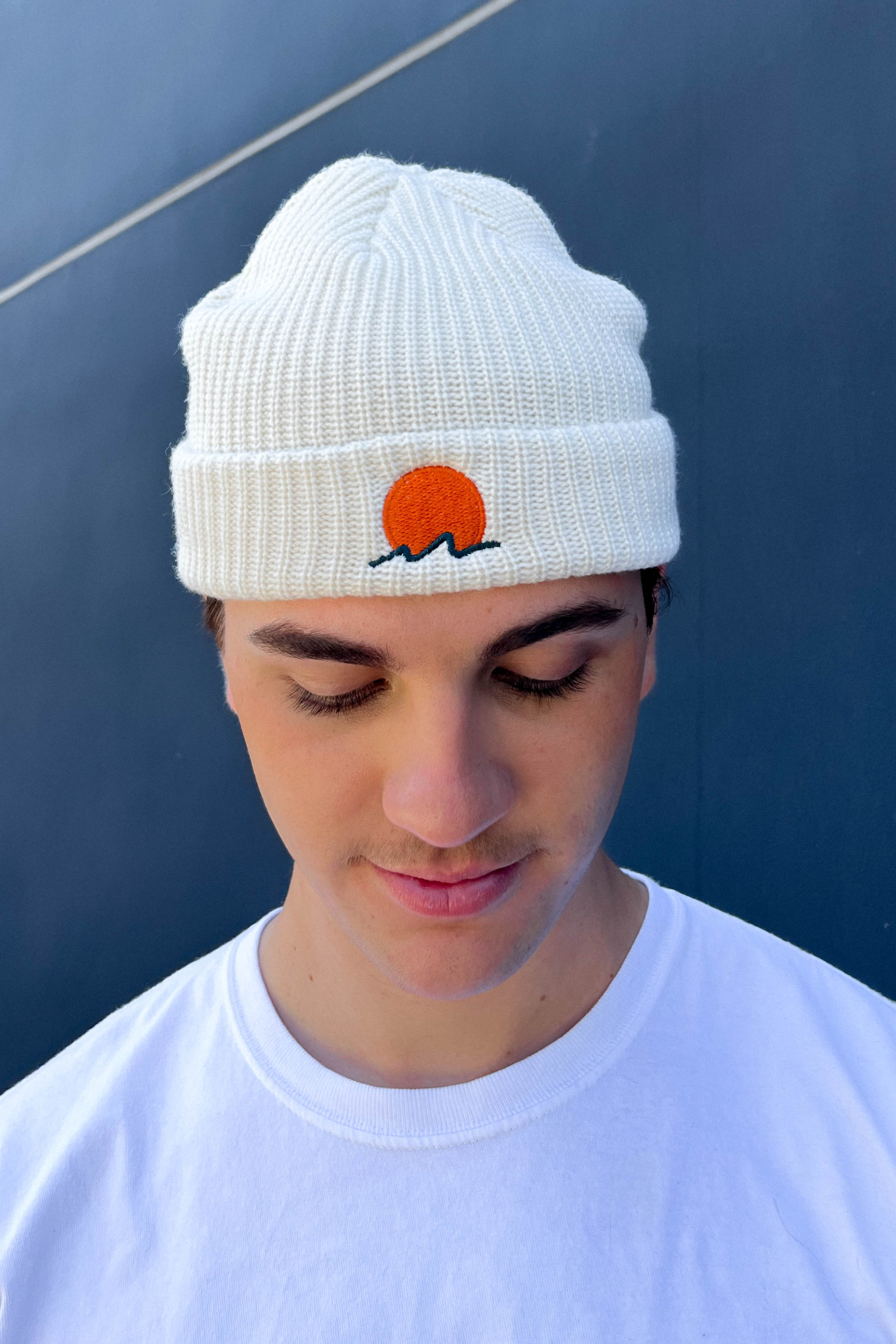 person wears a classic cuffed knit fisherman beanie featuring embroidered Rooted Swim sunset logo in a soft warm white color. high quality knit beanie