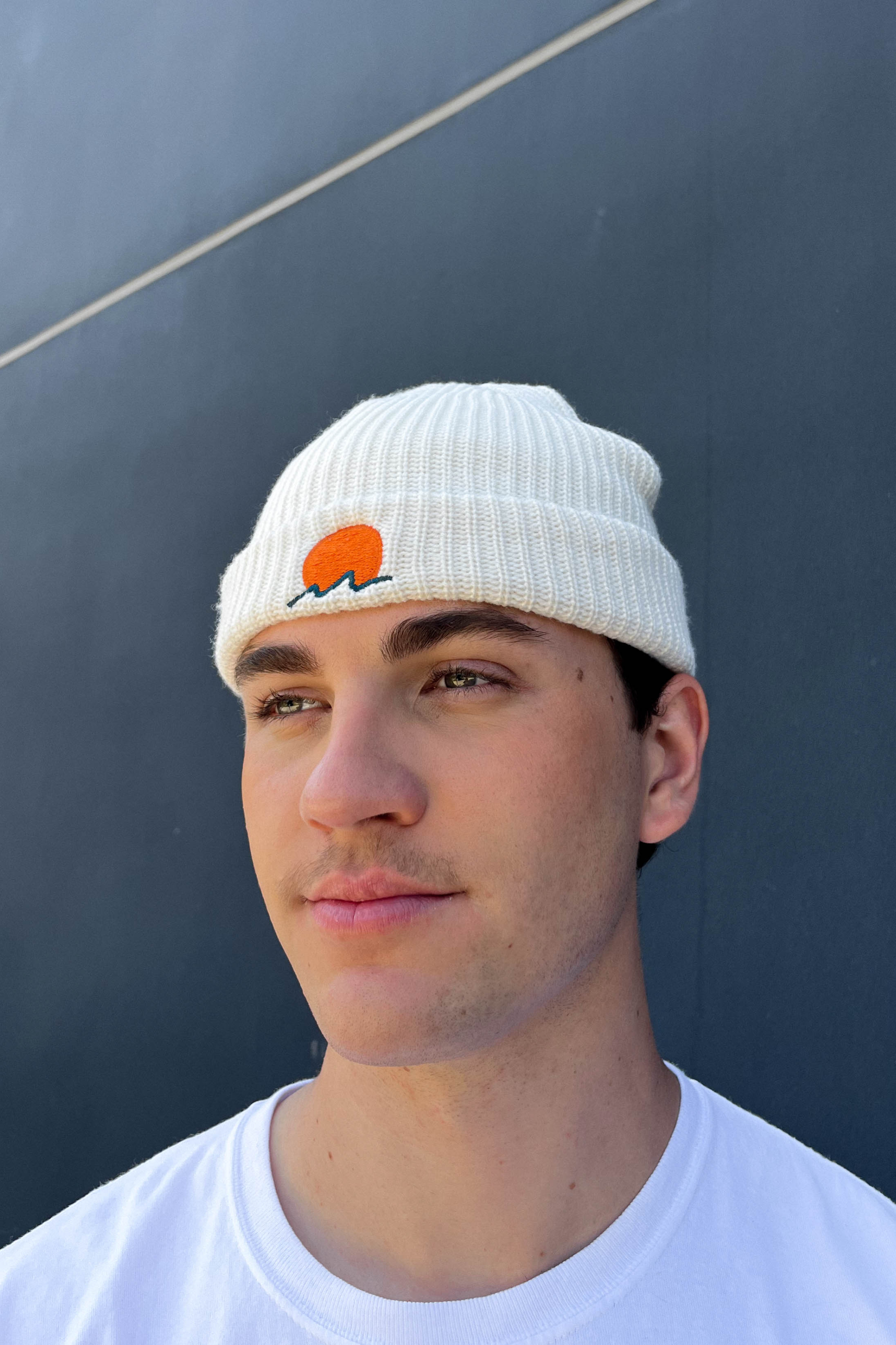 person wears a classic cuffed knit fisherman beanie featuring embroidered Rooted Swim sunset logo in a soft warm white color. high quality knit beanie