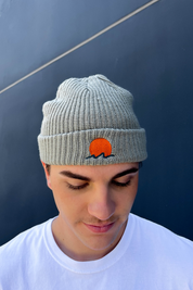 person wears classic cuffed knit fisherman beanie featuring embroidered Rooted Swim sunset logo in a soft green gray color. high quality knit beanie