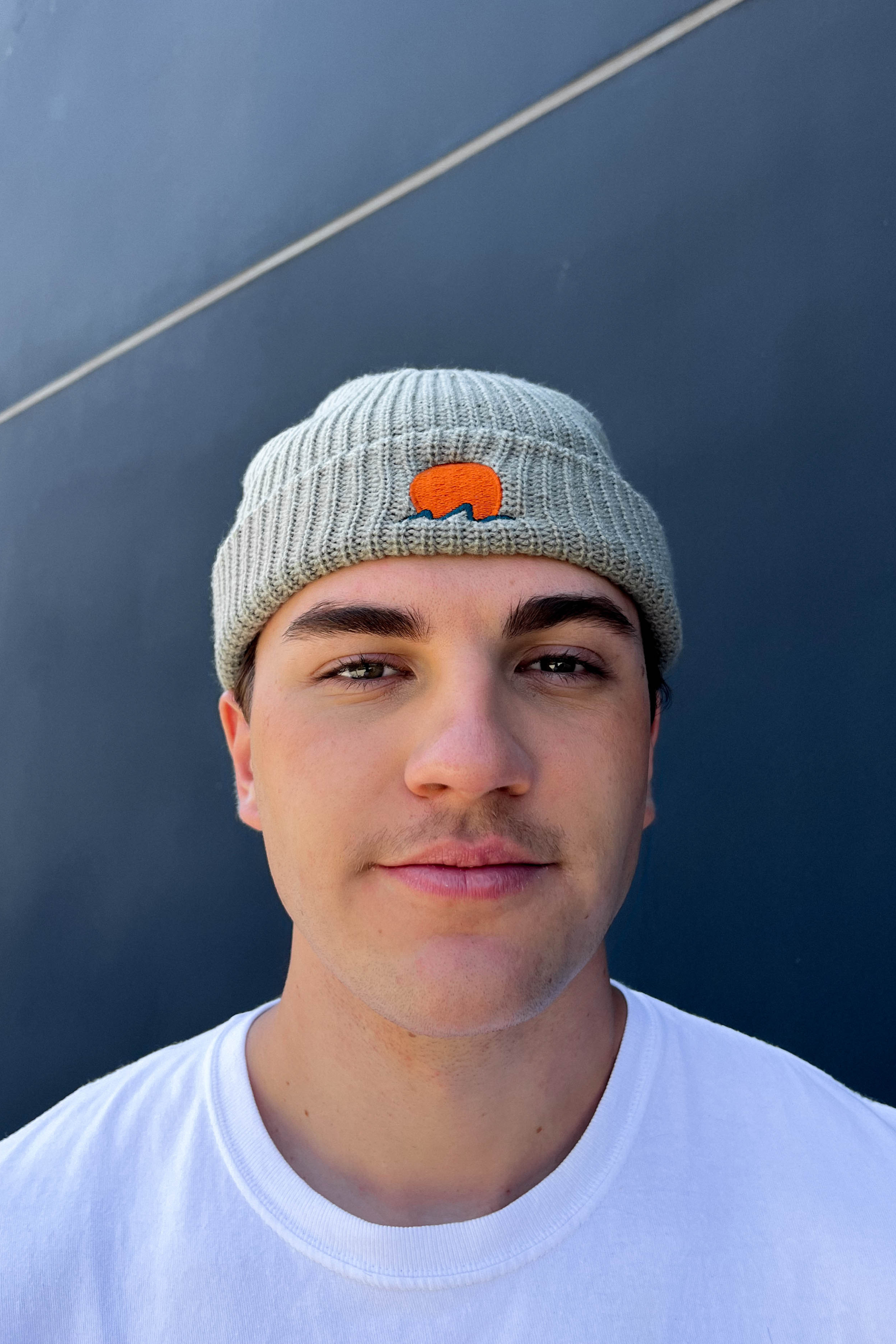person wears classic cuffed knit fisherman beanie featuring embroidered Rooted Swim sunset logo in a soft green gray color. high quality knit beanie
