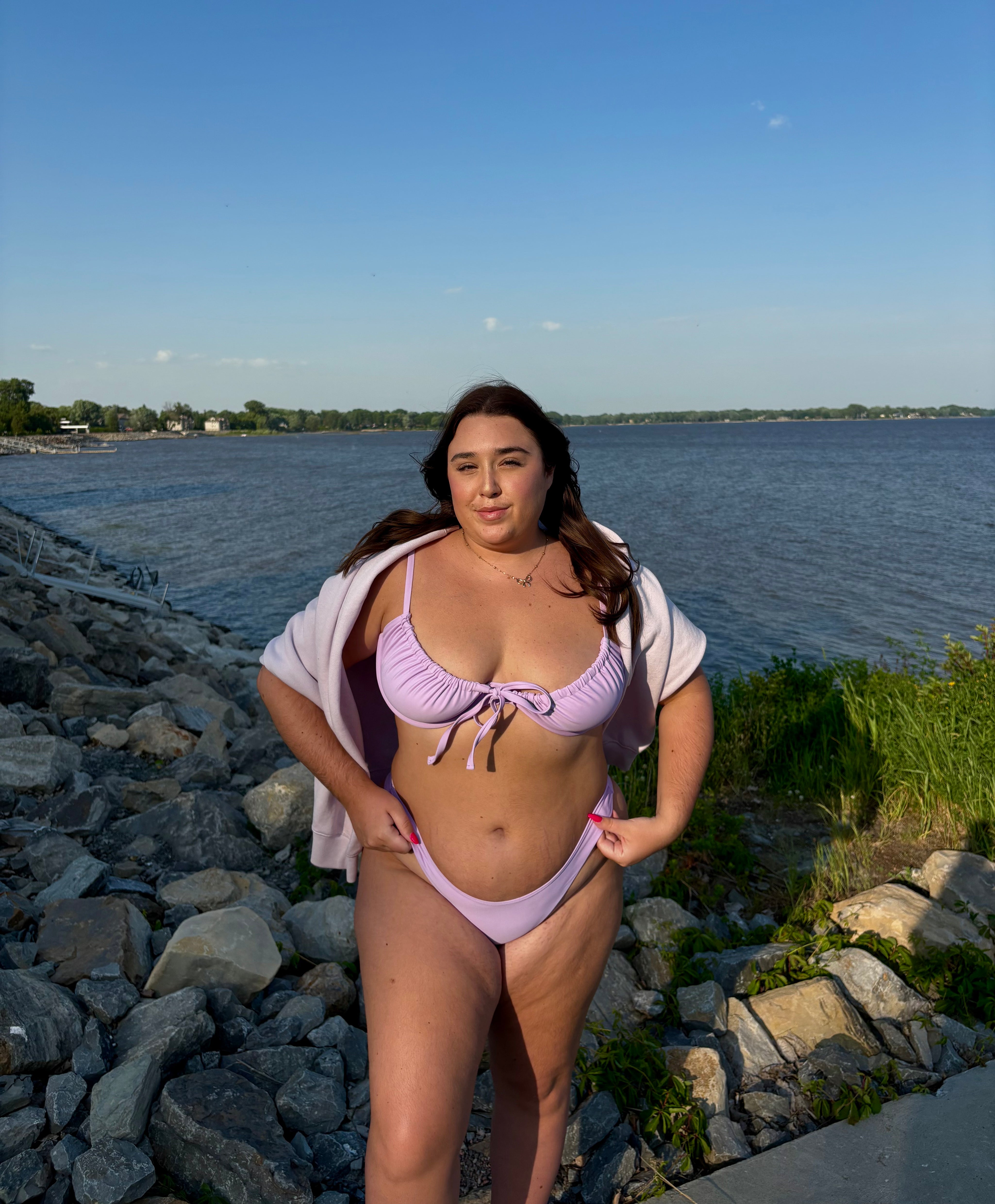 Woman in a light purple bikini standing by a body of water with a clear sky.