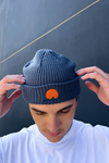 Person wearing classic cuffed knit fisherman beanie featuring embroidered Rooted Swim sunset logo in a blue gray color. high quality knit beanie