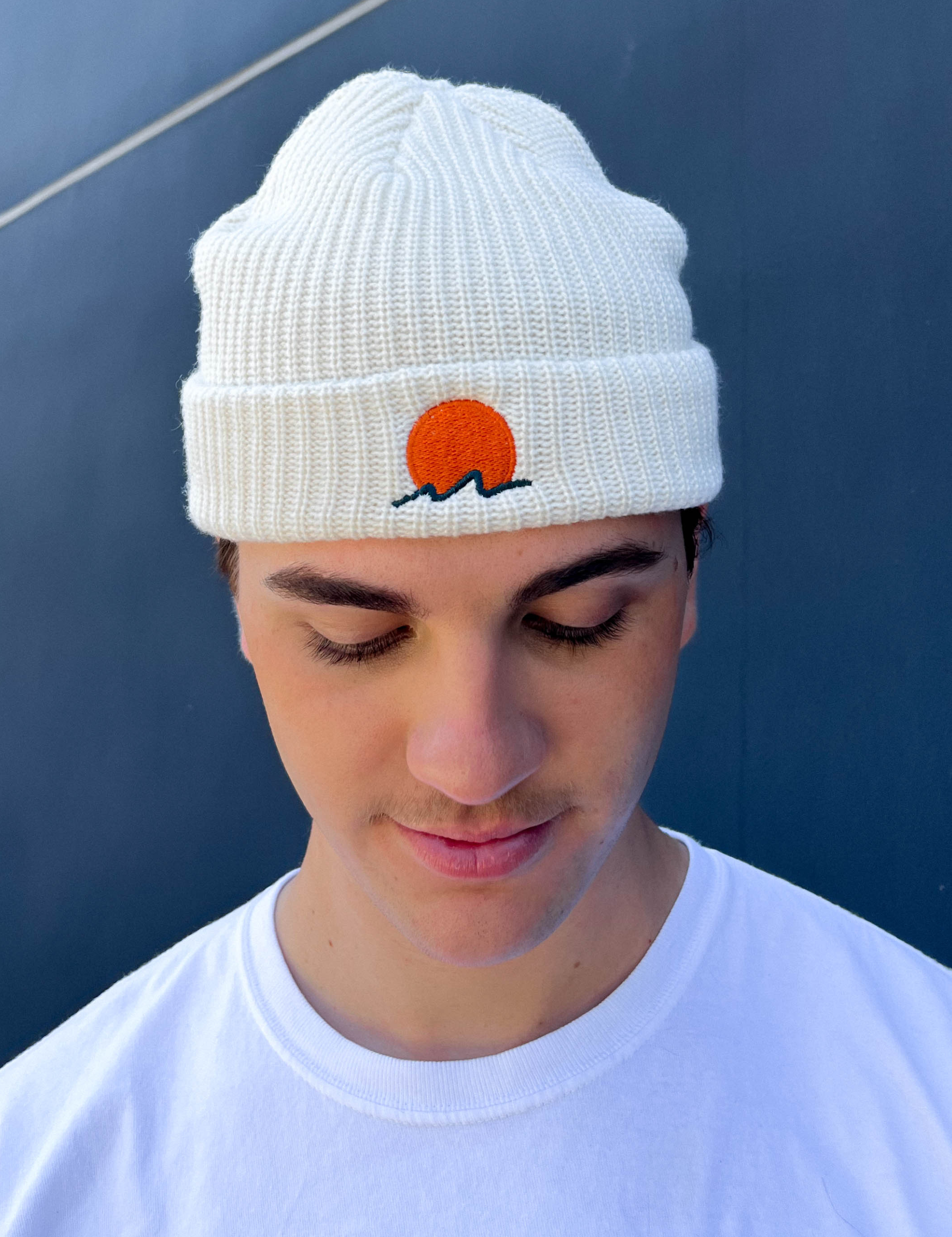 person wears a classic cuffed knit fisherman beanie featuring embroidered Rooted Swim sunset logo in a soft warm white color. high quality knit beanie