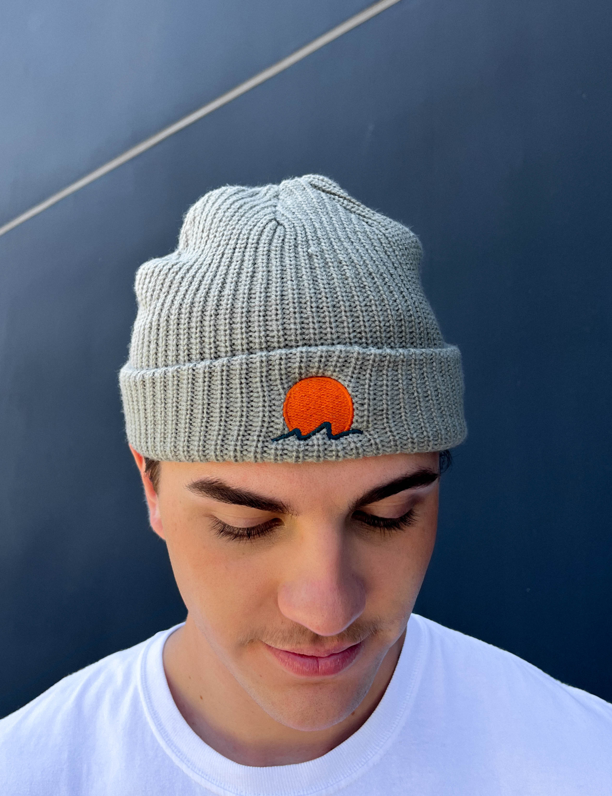 person wears classic cuffed knit fisherman beanie featuring embroidered Rooted Swim sunset logo in a soft green gray color. high quality knit beanie